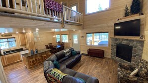 Une cabane spacieuse comprenant une chambre en mezzanine, un coin repas, un salon avec cheminée et une cuisine dotée de meubles en bois.