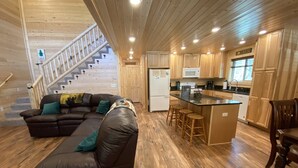 Un vaste espace de vie comprenant un canapé en cuir marron, un plancher en bois, une cuisine avec des meubles en bois, un îlot central doté de tabourets et un escalier menant aux étages supérieurs.
