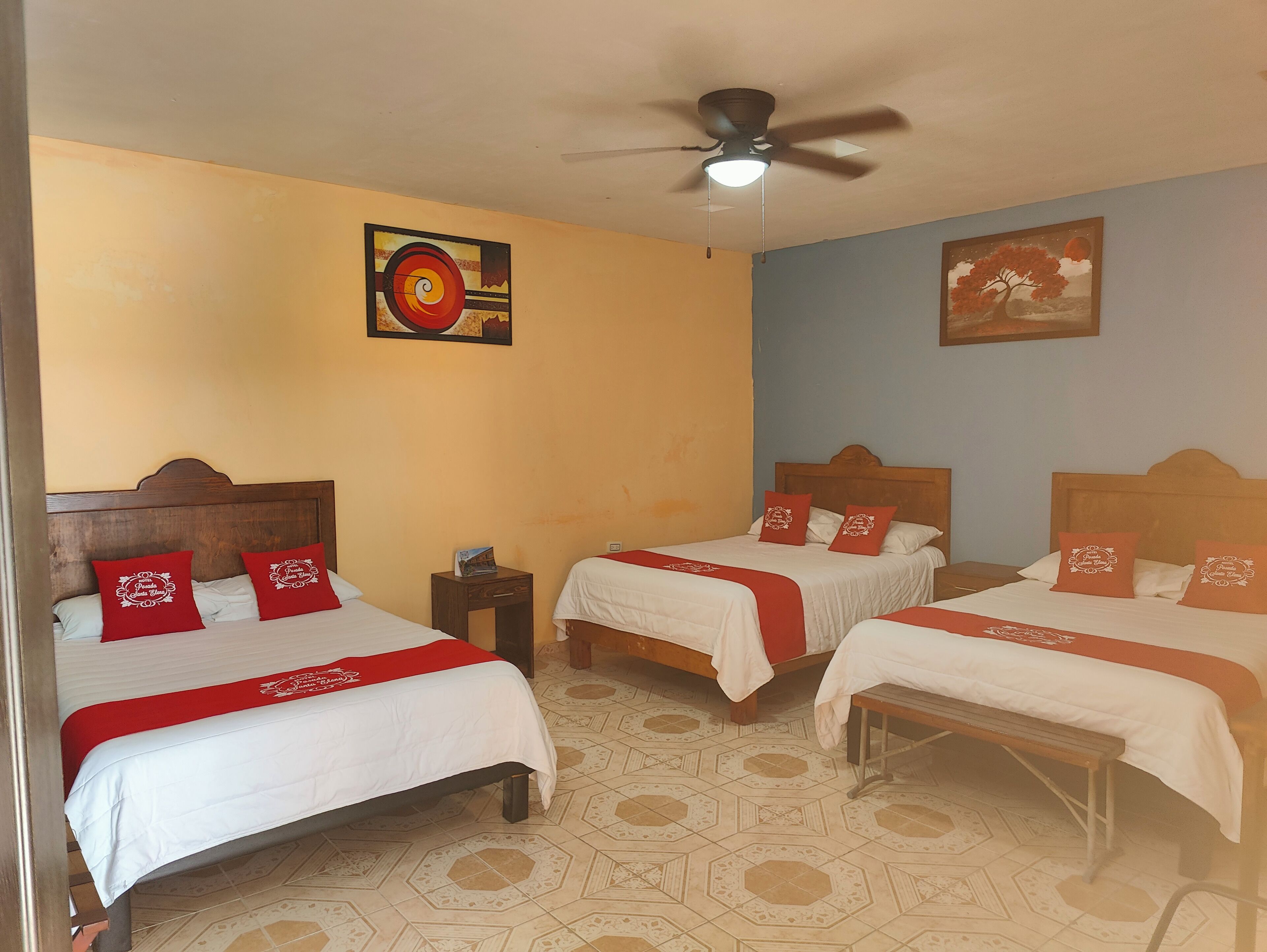 A hotel room with three beds, each with red pillows and white bedding. There is a ceiling fan, a wall clock, and two paintings.