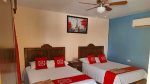 Ein Hotelzimmer mit zwei Betten, jeweils mit weißer Bettwäsche und roten Zierkissen. Es gibt einen Deckenventilator, ein gerahmtes Bild an der Wand und eine Klimaanlage.