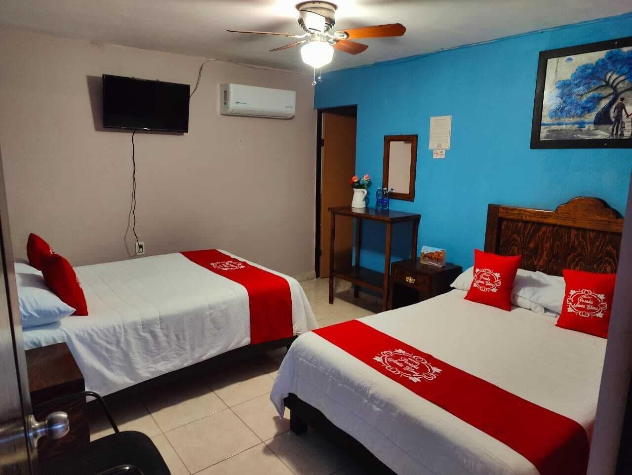 A hotel room with two beds, each with red and white bedding, a ceiling fan, a mounted TV, and a painting on the wall.