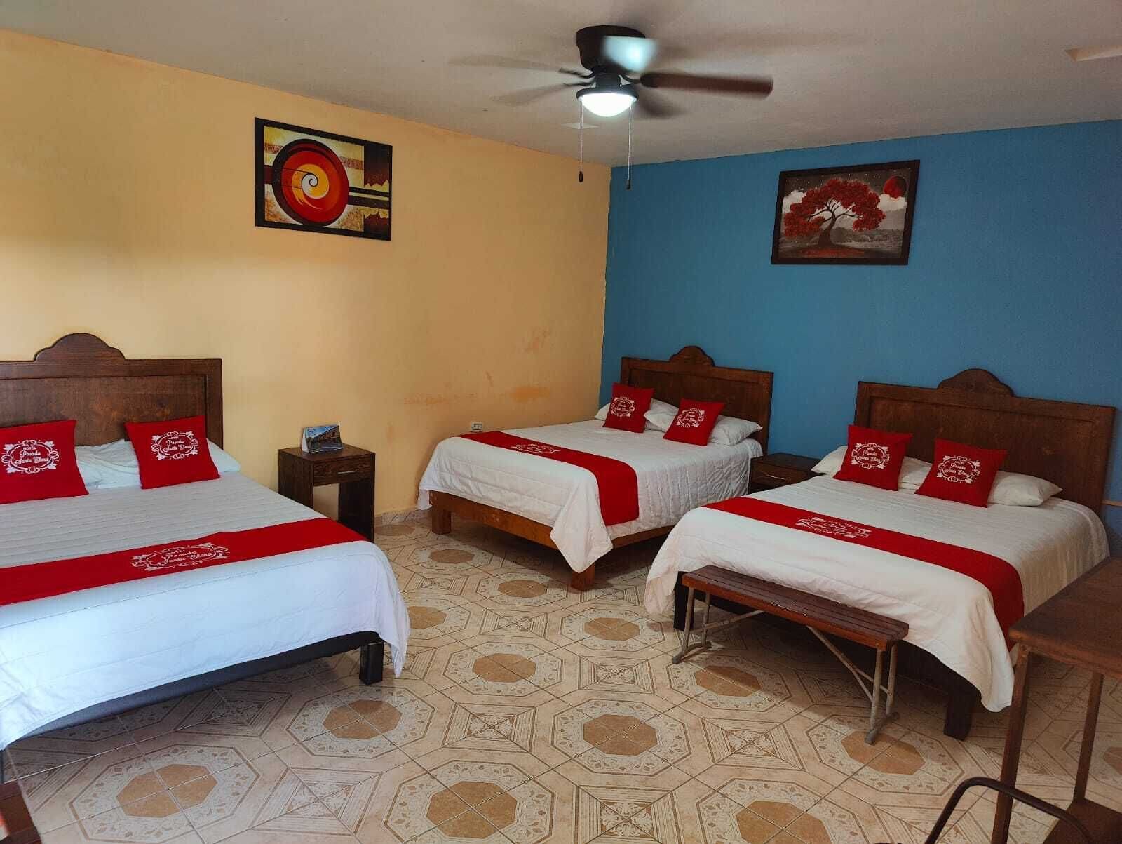 Habitación de hotel con tres camas, cada una con almohadas rojas y colcha blanca. Hay un ventilador de techo, una mesita y una pared con un cuadro.