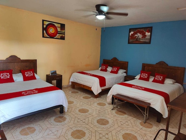 Habitación de hotel con tres camas, cada una con almohadas rojas y colcha blanca. Hay un ventilador de techo, una mesita y una pared con un cuadro.