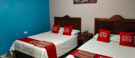 A hotel room with two beds, each with red pillows and a white bedspread. There is a painting on the wall above the beds, and an air conditioning unit mounted on the blue wall.