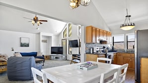 A spacious living area with a dining table, white chairs, a sofa, and a TV. There is a kitchen with wooden cabinets and a stone backsplash. - Lake View and close to Beach (LAKE ISABELLA)
