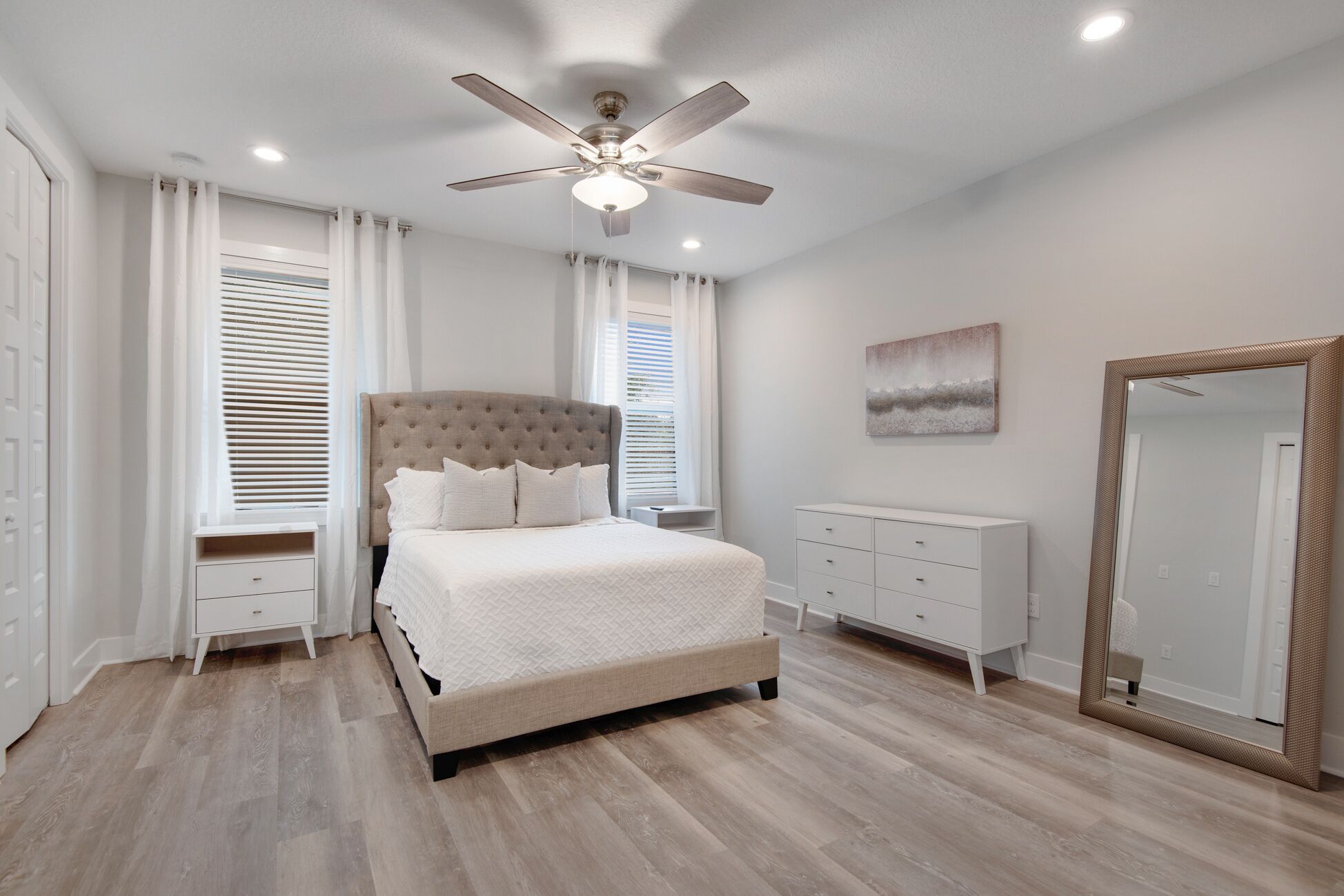 Une chambre à coucher comprenant un lit, un ventilateur de plafond, une commode, un miroir et une fenêtre avec des stores.