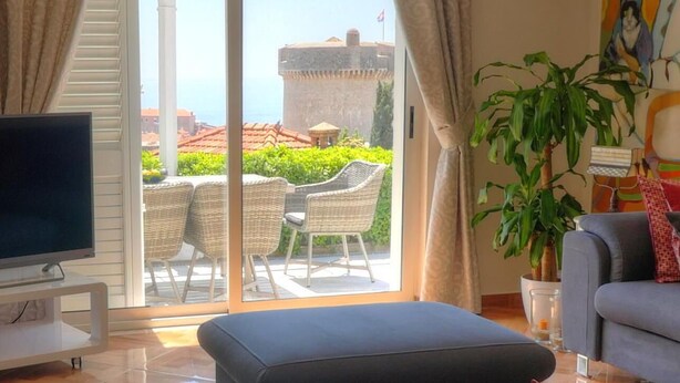 A living room with a TV, a sofa, a potted plant, and a view of a building and greenery through a sliding glass door.