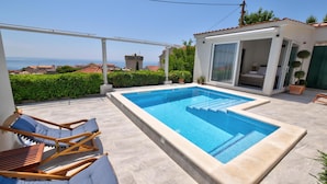 A rectangular swimming pool with a clear blue water, surrounded by a light-colored deck. There are two lounge chairs with blue cushions and a wooden table. The pool is adjacent to a building with a glass door and a small balcony.
