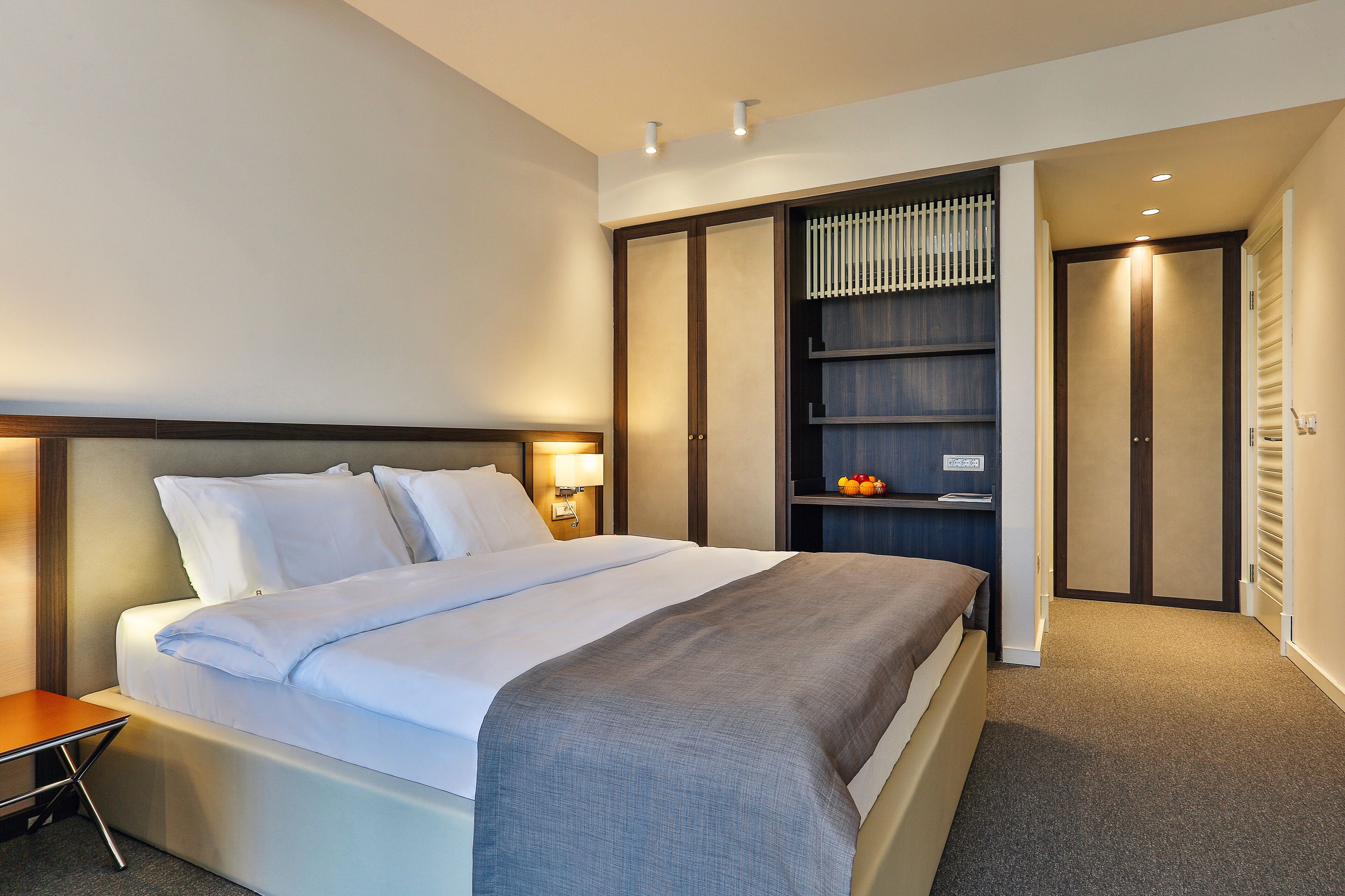 A modern hotel room with a large bed, a bedside table, a wardrobe, and a shelf with fruits.