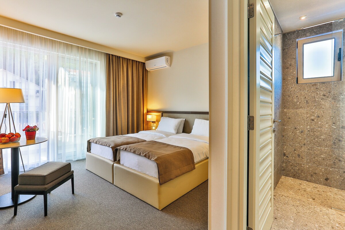 Habitación de hotel con cama, mesita de noche, lámpara y vistas al exterior por la ventana.