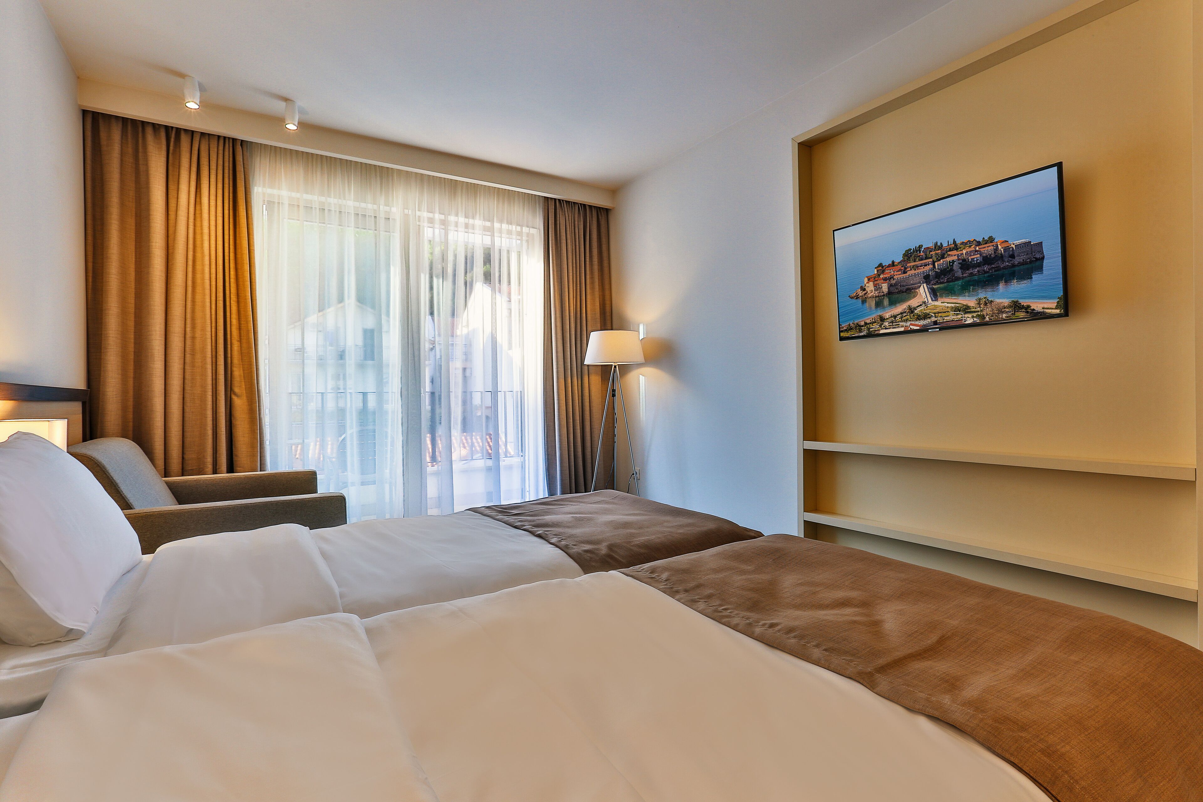 Habitación de hotel con dos camas, un ventanal con cortinas, una lámpara de pie y un cuadro colgado en la pared.