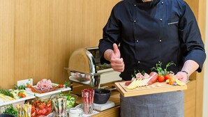 En kok i sort uniform og hvid kokkehue står i et køkken med forskellige madvarer og en køledisk.