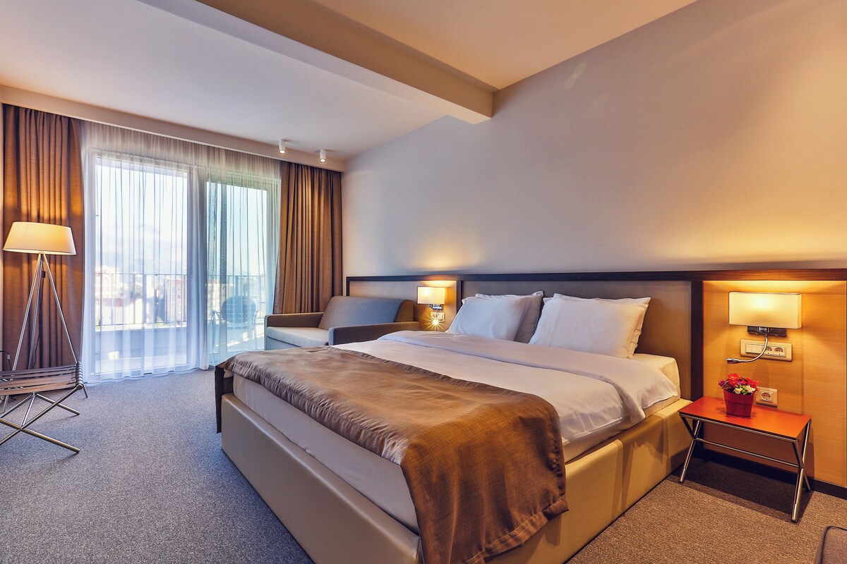 Habitación de hotel con una cama grande, dos mesitas de noche con lámparas, una mesita pequeña con un florero y vistas a la ciudad a través de las ventanas.