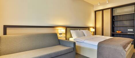 A hotel room with a grey sofa, a bed with a grey blanket, a nightstand with a lamp, and a wardrobe with a mirror.