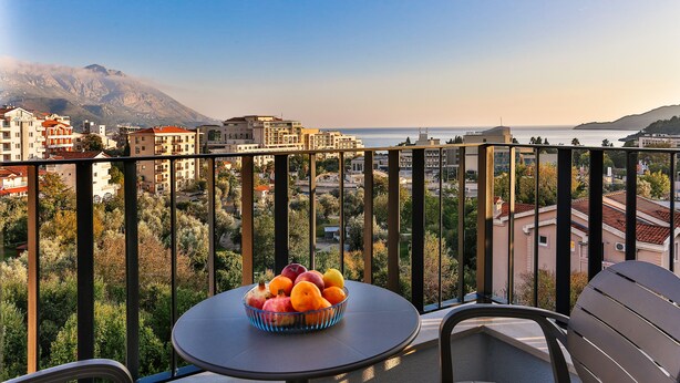 A balcony with a table and chairs, a bowl of fruit, and a view of the sea and buildings. - Hotel Bono (Becici)