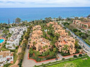 A coastal residential area with multiple buildings, including a prominent pinkish complex with a central courtyard surrounded by palm trees.