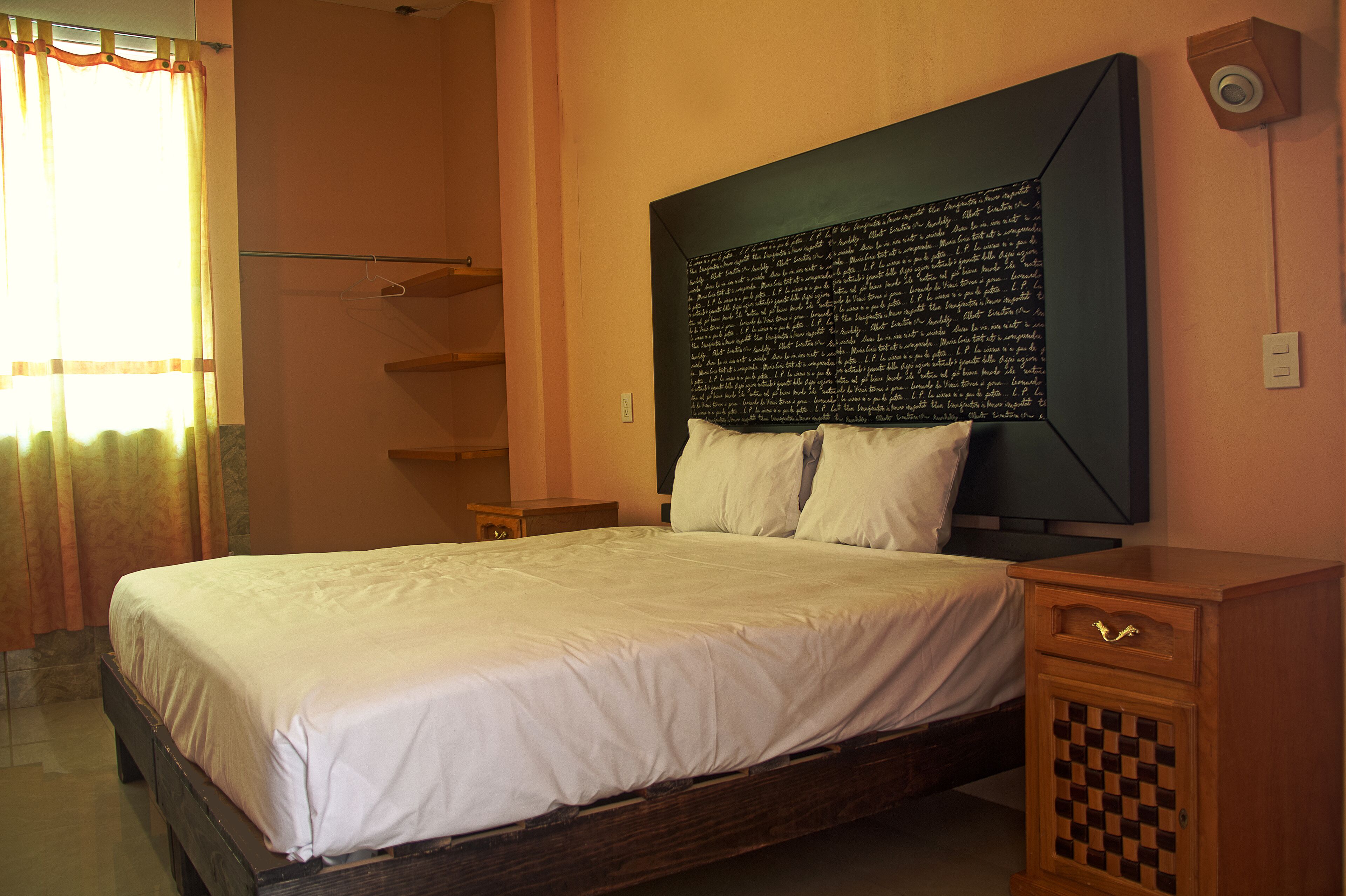 Uma cama bem arrumada com cabeceira com texto, uma mesa de cabeceira de madeira com gaveta, e uma câmera de segurança instalada na parede.