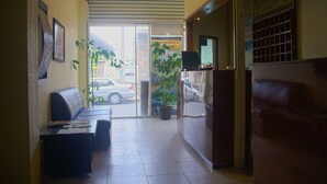 Un espace d’accueil d’hôtel comprenant un comptoir de réception, un coin salon avec un canapé et des fauteuils, des plantes en pot et une vue sur la rue.