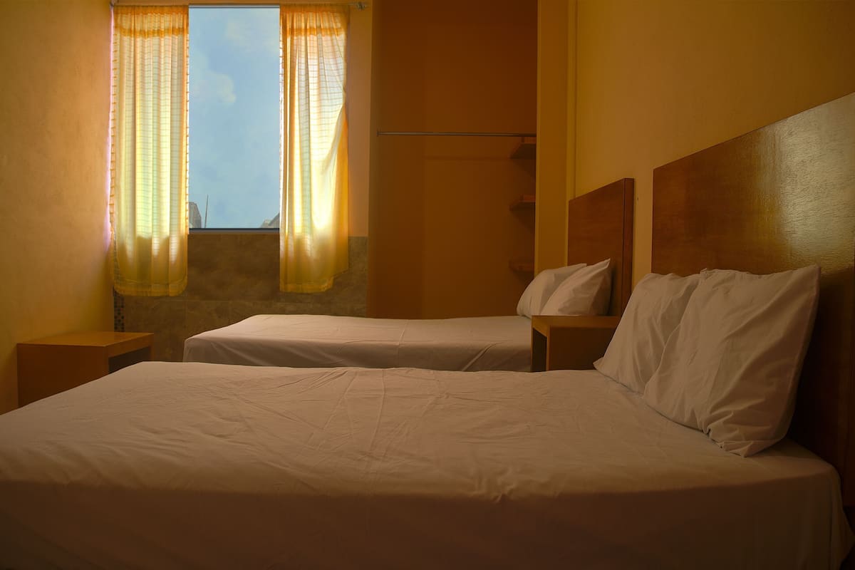 Ein Hotelzimmer mit zwei Betten, einem Holzkopfstück, einem Fenster mit Vorhängen und einer Steinwand.