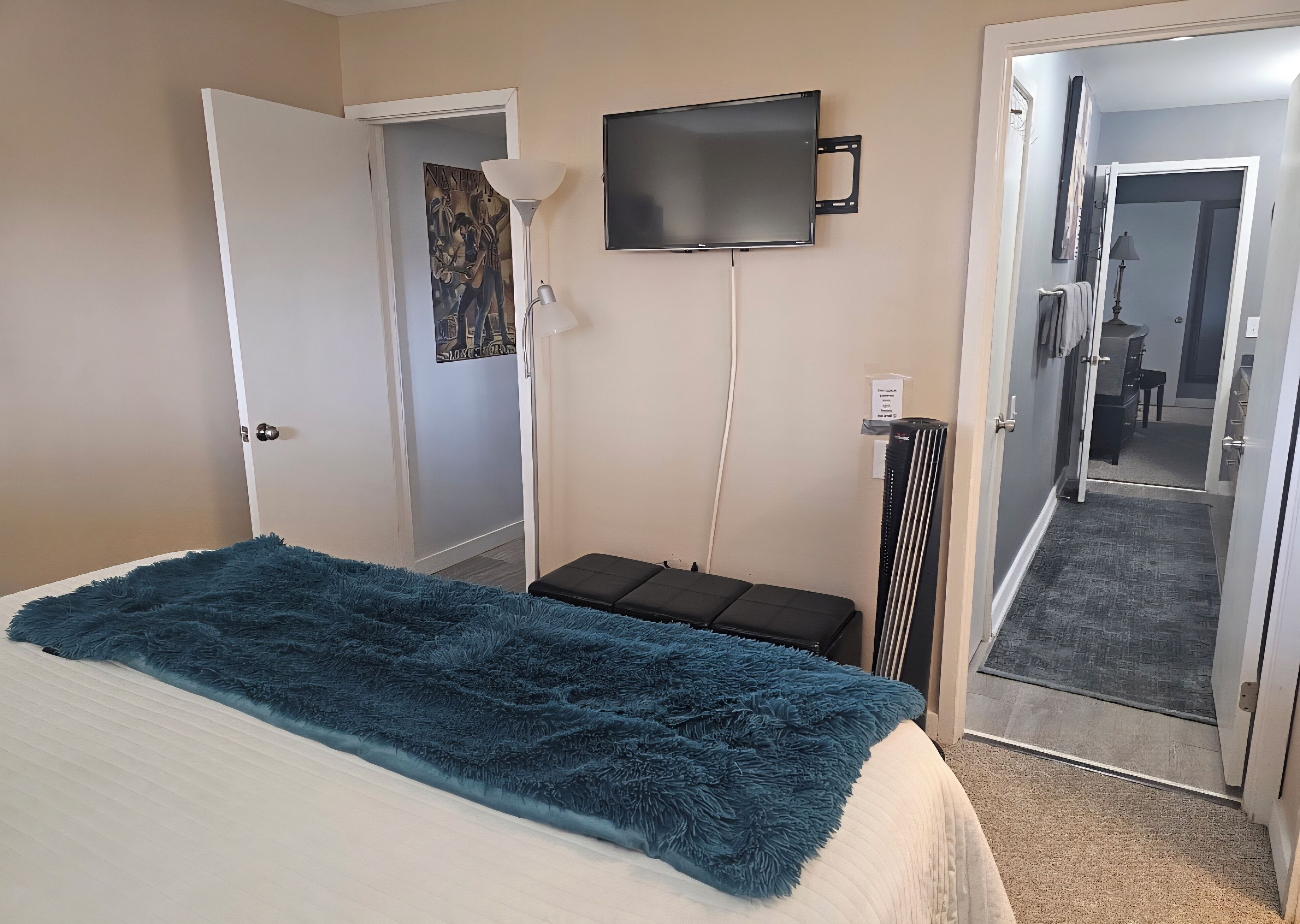 A hotel room with a bed, a TV mounted on the wall, a door leading to a hallway, and a small seating area with a bench.