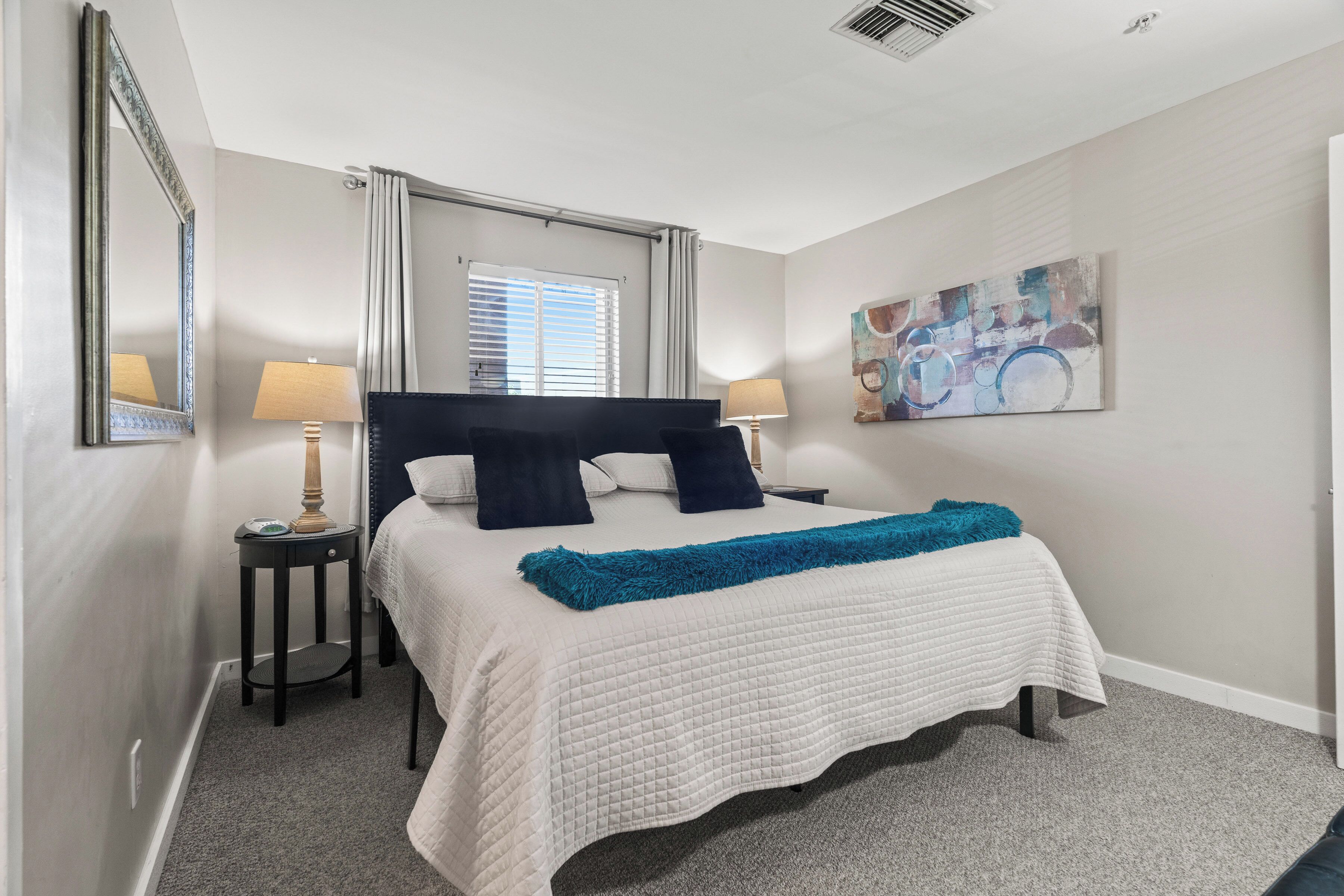 A neatly arranged bedroom with a large bed, two bedside tables with lamps, a window with blinds, and a wall-mounted abstract painting.