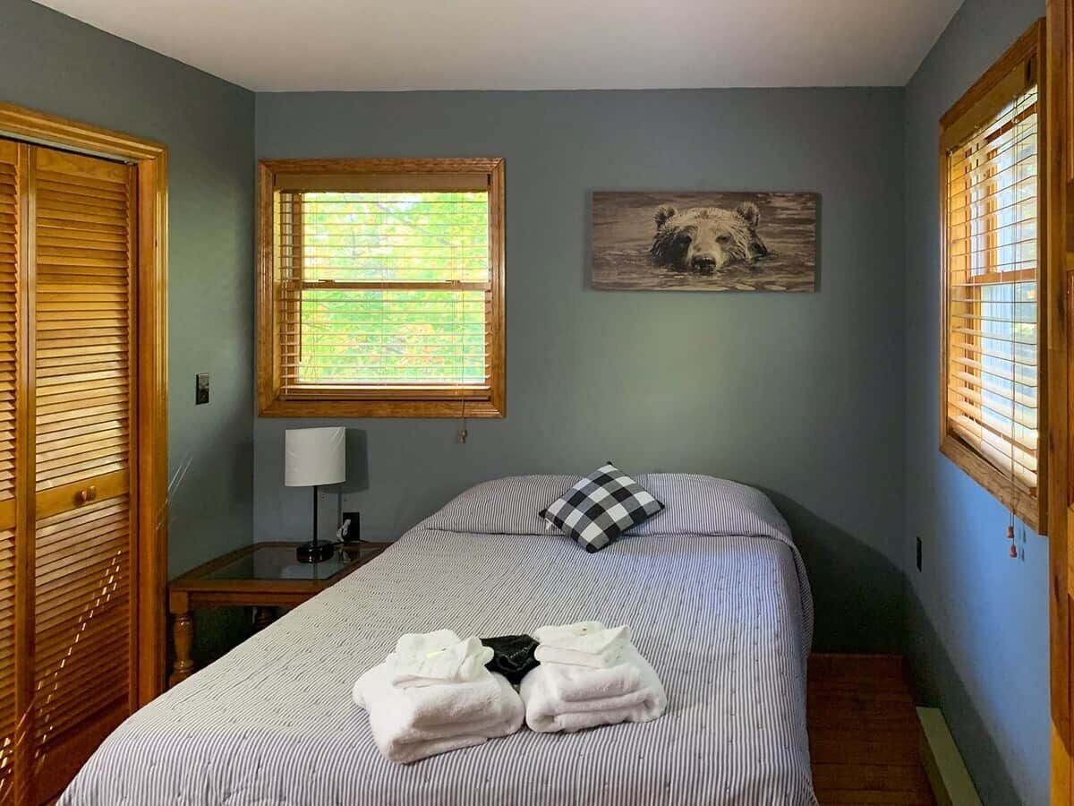A bedroom with a bed, a bedside table, a lamp, a window with blinds, and a wall-mounted picture.