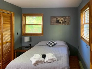 Une chambre à coucher comprenant un lit, une table de chevet, une lampe, une fenêtre avec des stores et un tableau accroché au mur.