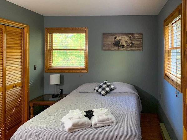 A bedroom with a bed, a bedside table, a lamp, a window with blinds, and a wall-mounted picture.