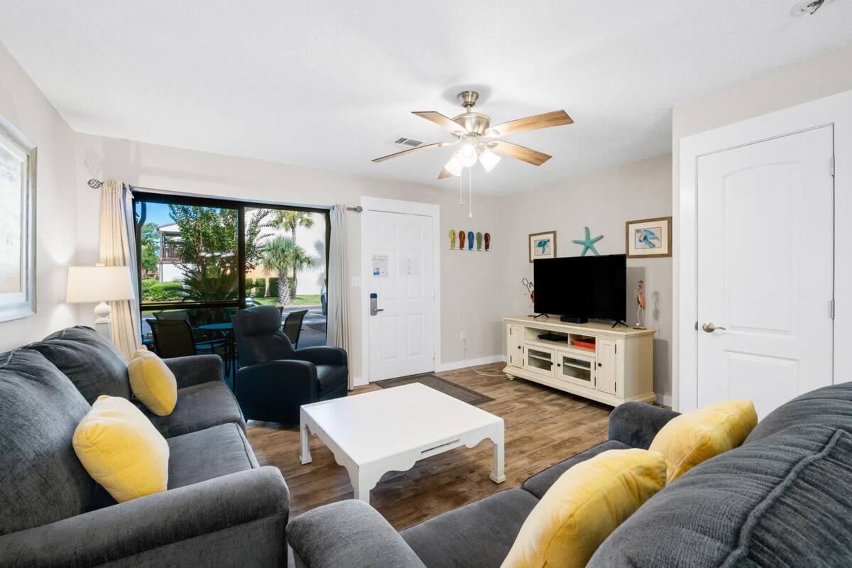 Una sala de estar con un ventilador de techo, un televisor sobre un mueble blanco y un amplio ventanal con vistas a palmeras.