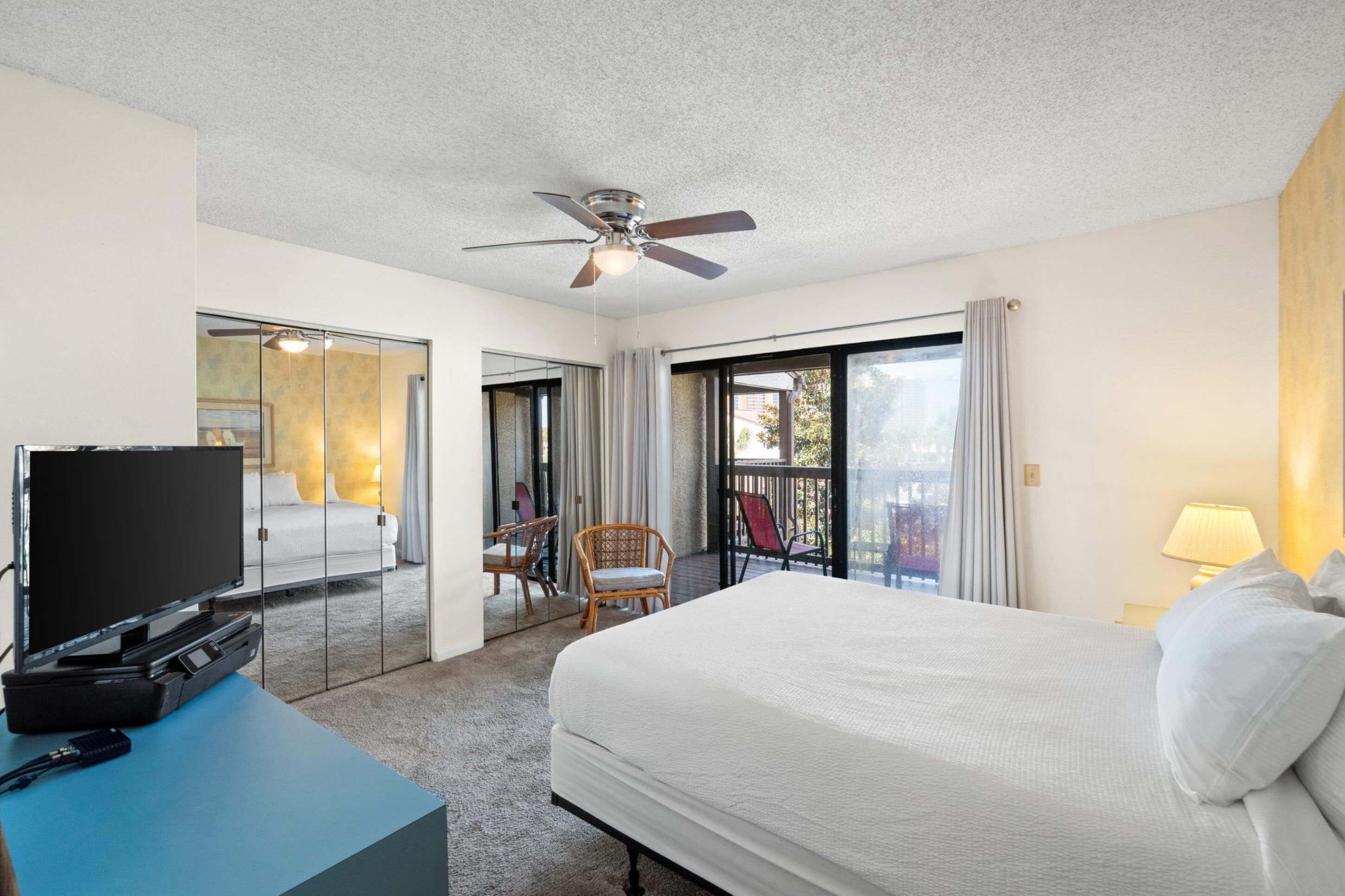A hotel room with a large bed, a ceiling fan, a television on a stand, a balcony with a chair, and a sliding glass door leading to a balcony.