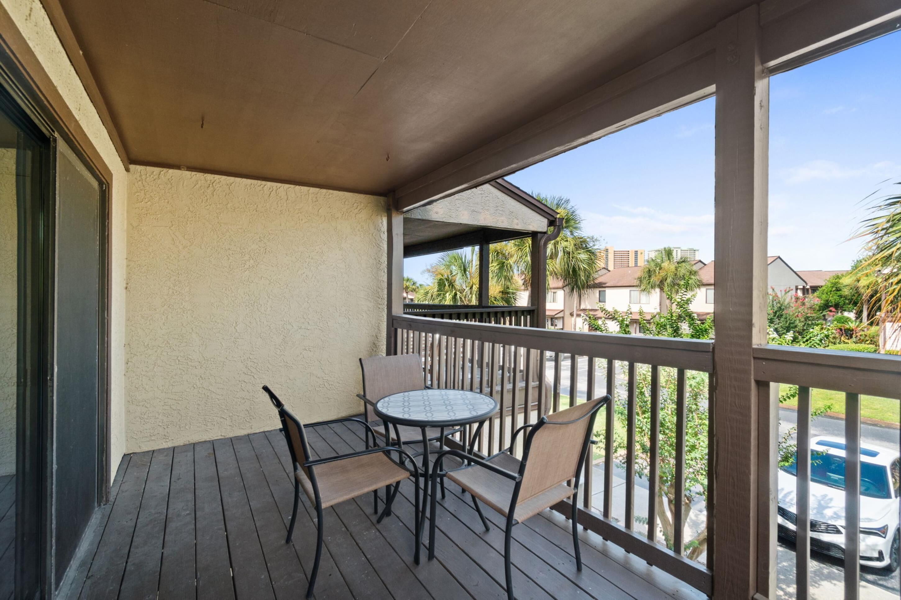 A covered outdoor area with a wooden deck, a round table with four chairs, and a view of palm trees and houses in the background.