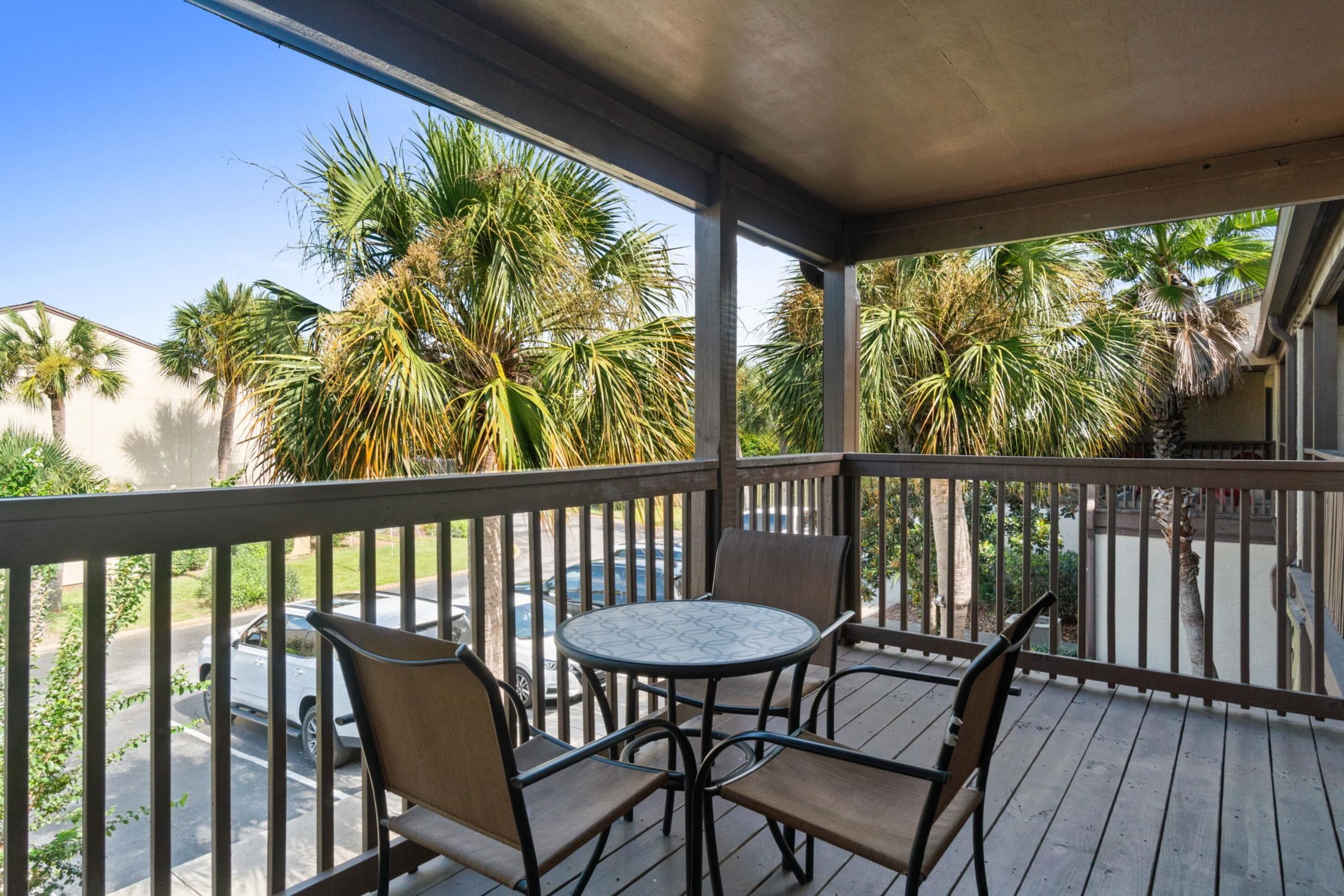 A covered outdoor area with a round table and four chairs, surrounded by a wooden deck and palm trees.