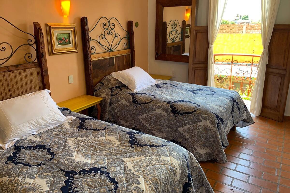 A bedroom with two beds, a wooden headboard, a bedside table, a wall-mounted mirror, and a window with curtains.