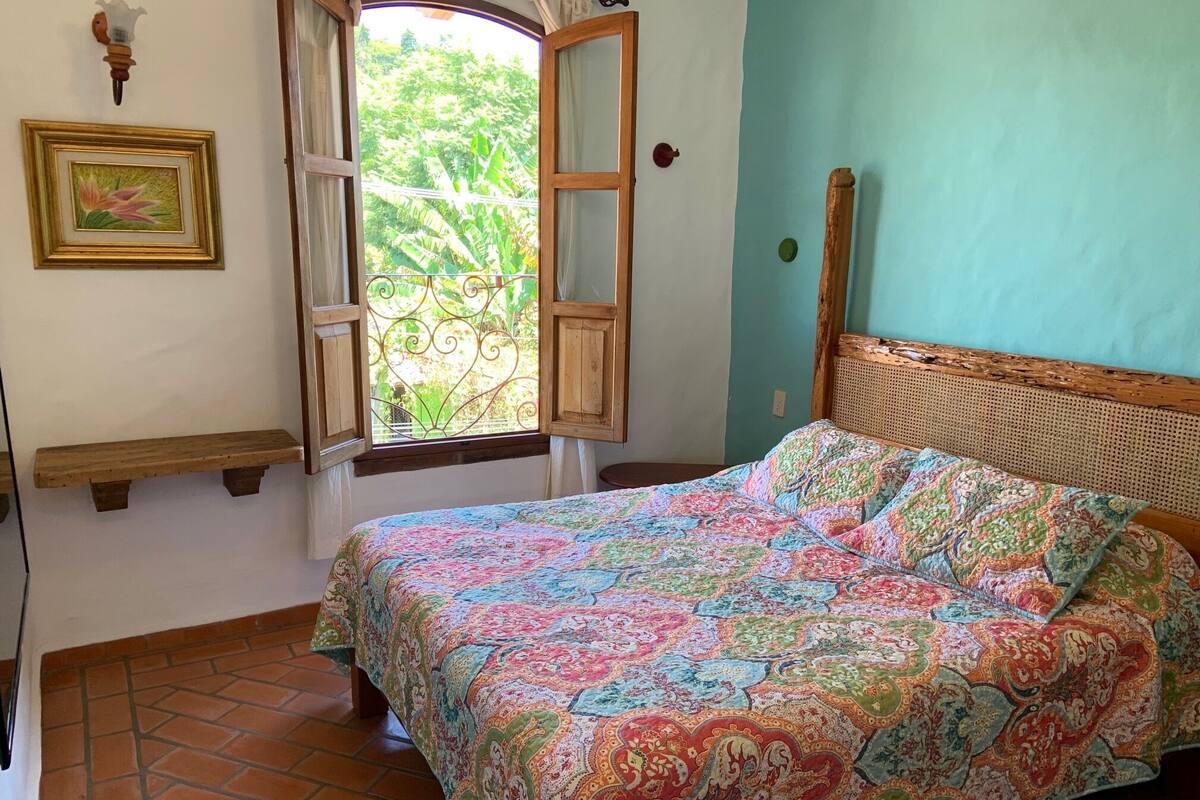 A bedroom with a bed, a window with open shutters, a picture frame, and a wooden bench.