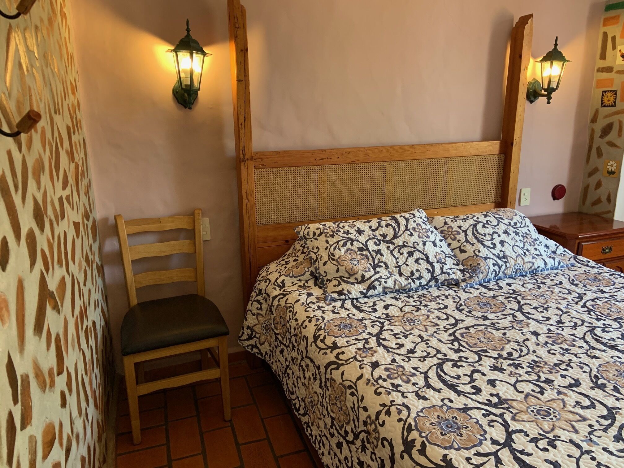 A bedroom with a wooden bed, patterned bedding, a wooden chair, and two wall-mounted lamps.