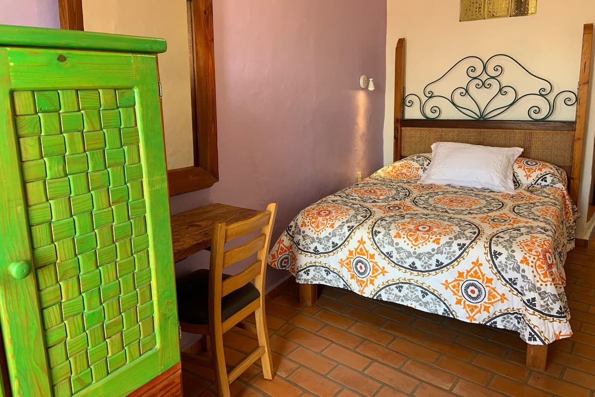 A bedroom with a bed, a wooden chair, a green door, and a wall-mounted light fixture.