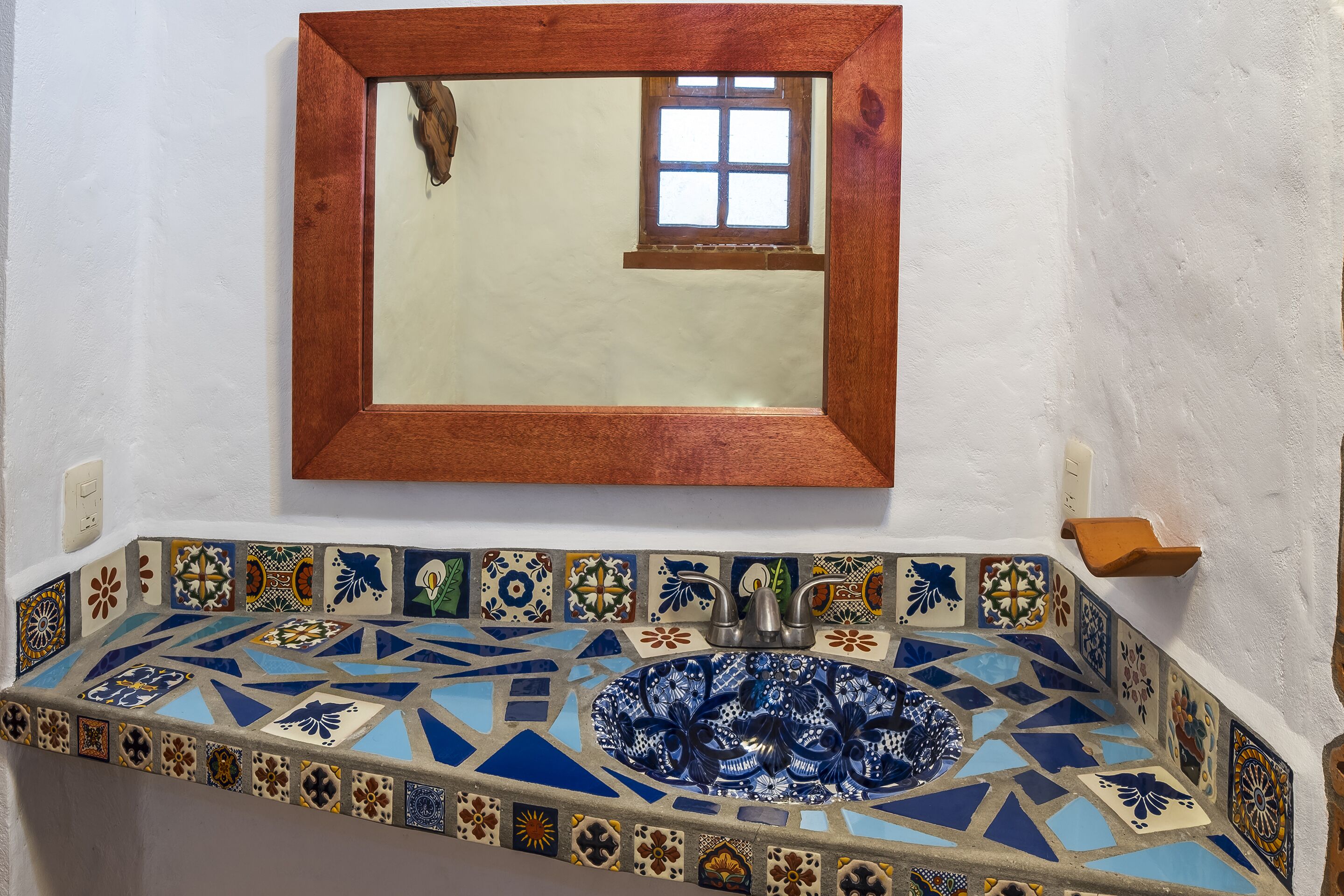Un baño con un lavamanos de mosaico, un espejo con marco de madera y una pequeña ventana.