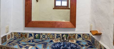 Une salle de bain avec un lavabo recouvert de mosaïques, un miroir encadré en bois et une petite fenêtre.