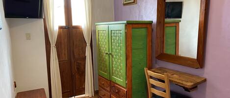A room with a wooden wardrobe, a small wooden table, a chair, a television mounted on the wall, and a window with curtains.