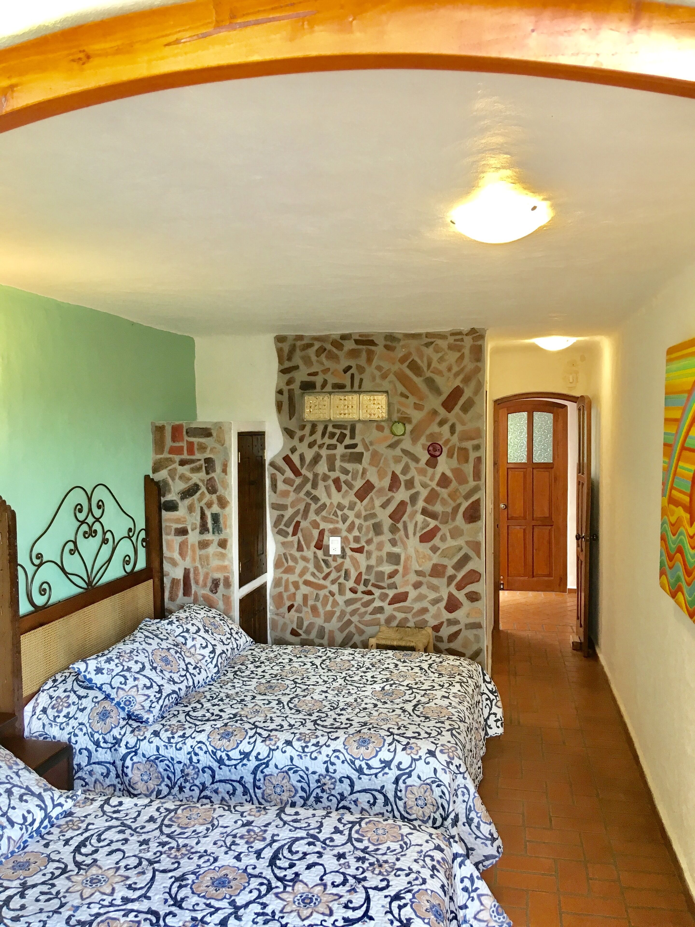 Un dormitorio con pared de piedra, cama con ropa de cama estampada, cabecero de madera y una puerta que da a otra habitación.