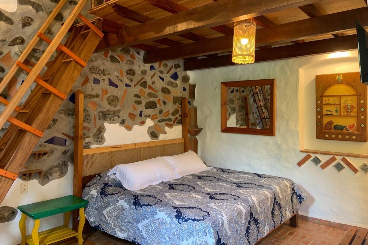 A bedroom with a stone wall, a wooden ladder, a bed with patterned bedding, a green bench, and a hanging light fixture.