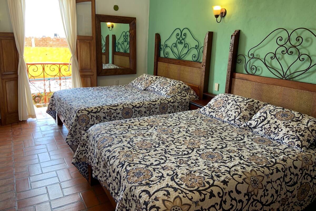 A room with two beds, a wooden door, a balcony with a metal railing, and a wall-mounted light fixture.