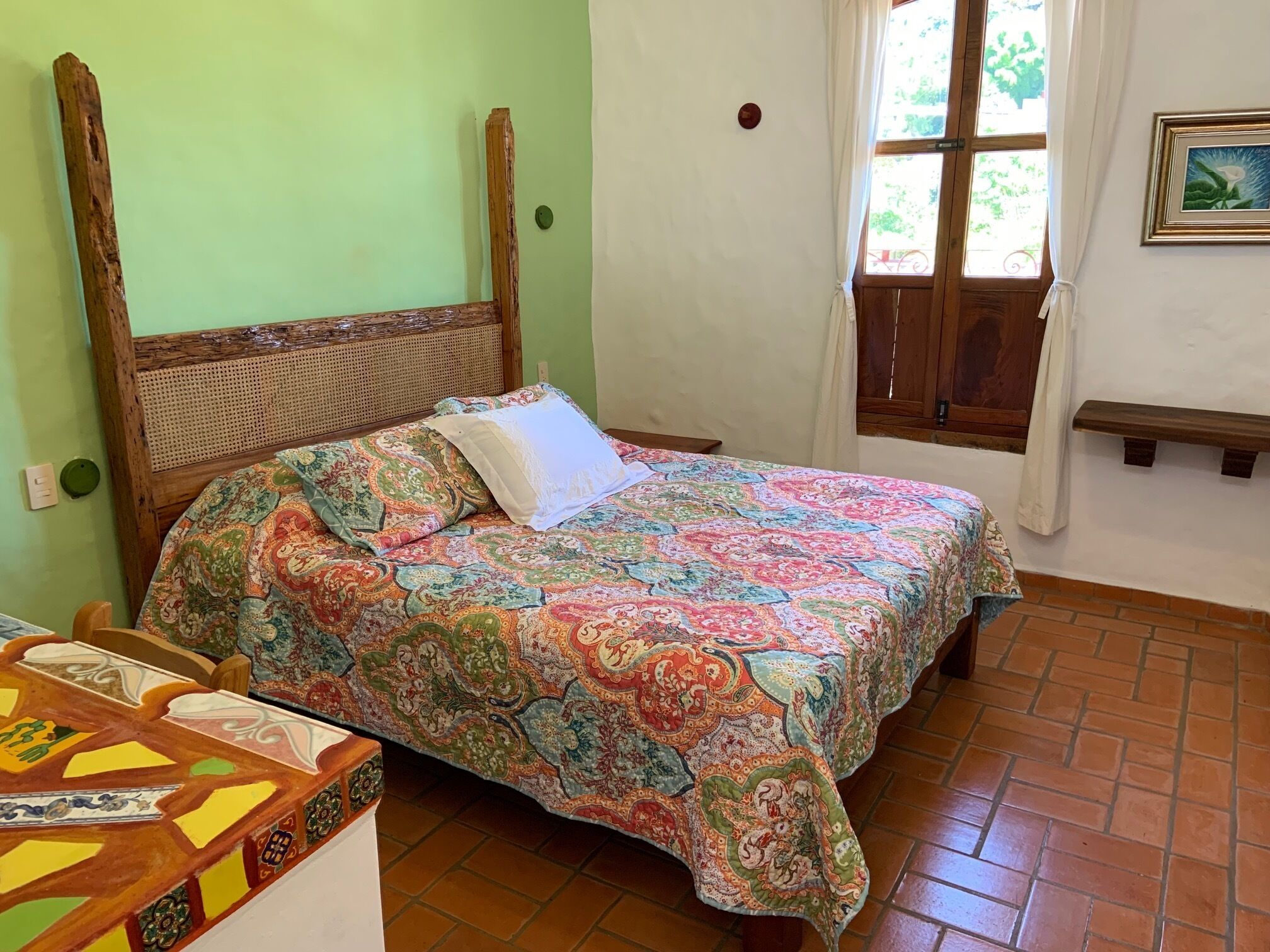 A room with a bed, a wooden headboard, a window with curtains, a bench, and a wall-mounted picture.