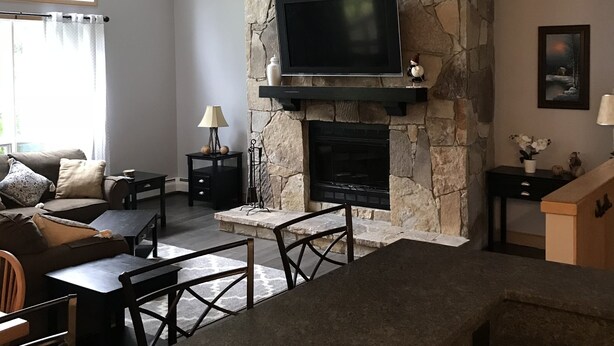 A living room with a stone fireplace, a television mounted above it, a ceiling fan, and a dining area with a table and chairs. - My Snow Slopeside Ski On/Off 4 BR Condo (Dover)