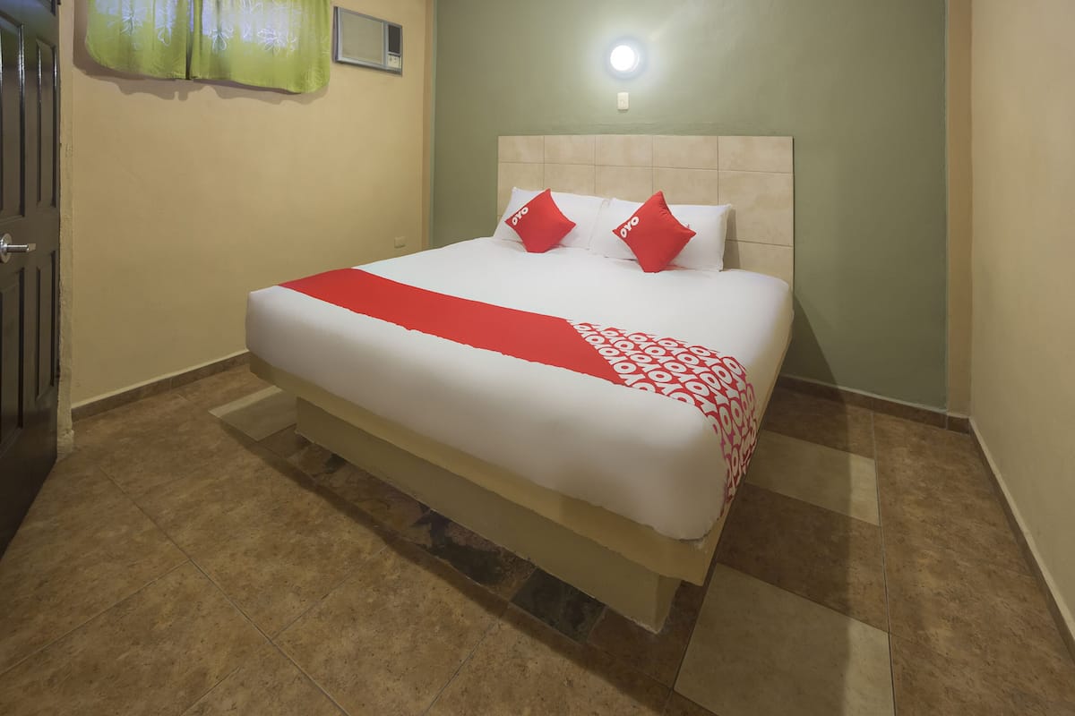 A neatly made bed with a red and white bedspread and two red pillows, a light-colored headboard, and a green wall with a window covered by a floral curtain.