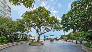 A spacious outdoor area with a large tree, seating arrangements, and a view of a building in the background.