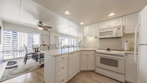 A modern kitchen with white cabinets, a microwave, stove, and a refrigerator. There is a dining area with a table and chairs, and a large window providing a view of the city.