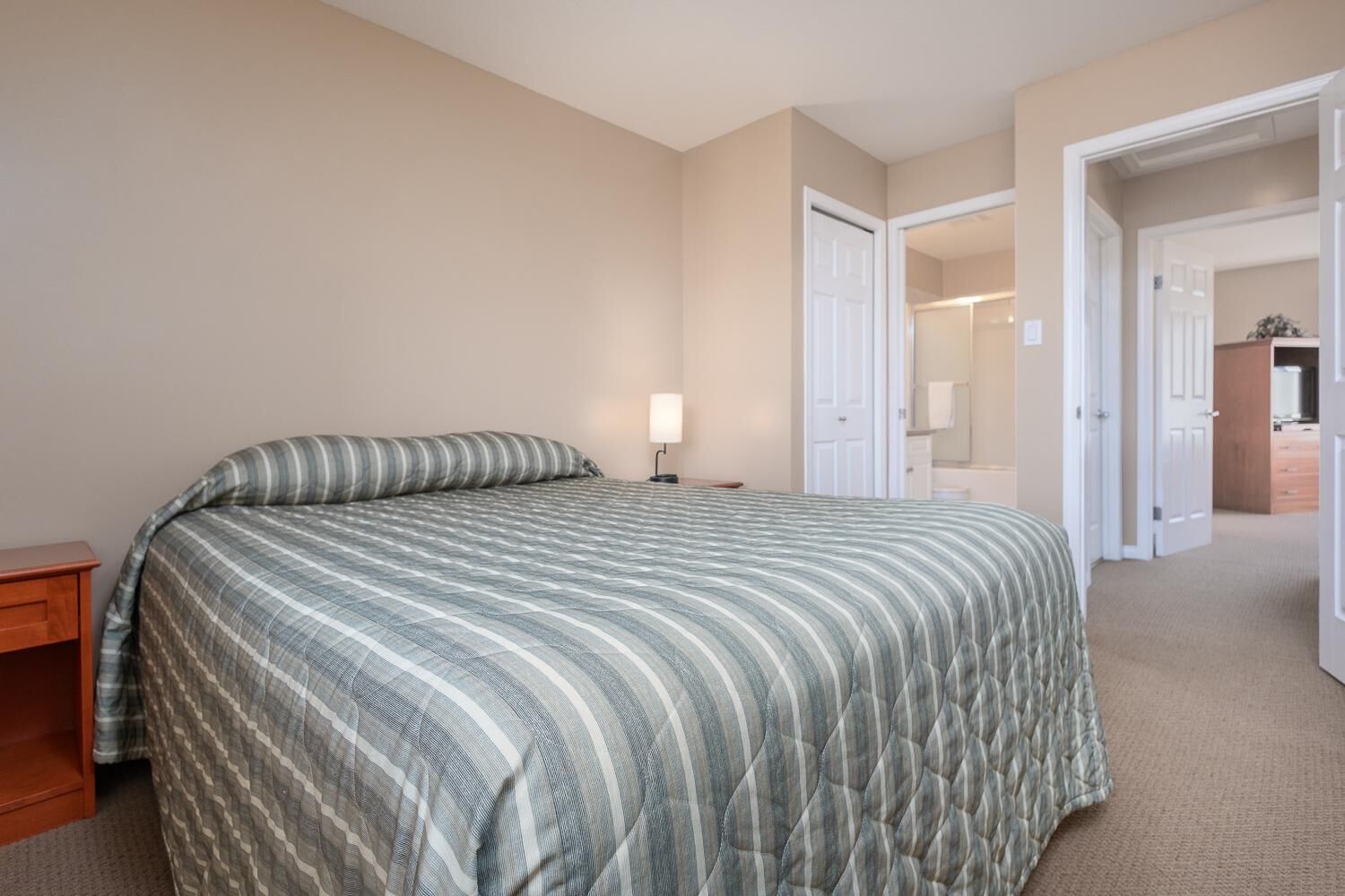 A bedroom with a large bed, a bedside table, a lamp, and a door leading to another room.