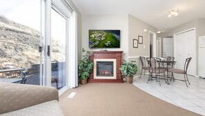 Una sala de estar con chimenea, un televisor montado sobre ella, una zona de comedor con sillas y una mesa, y un ventanal con vistas a una colina.