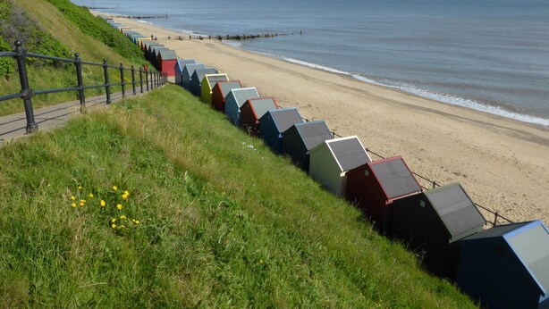 Una playa con cabañas coloridas alineadas a lo largo de la orilla, un área verde con una valla y el océano al fondo.